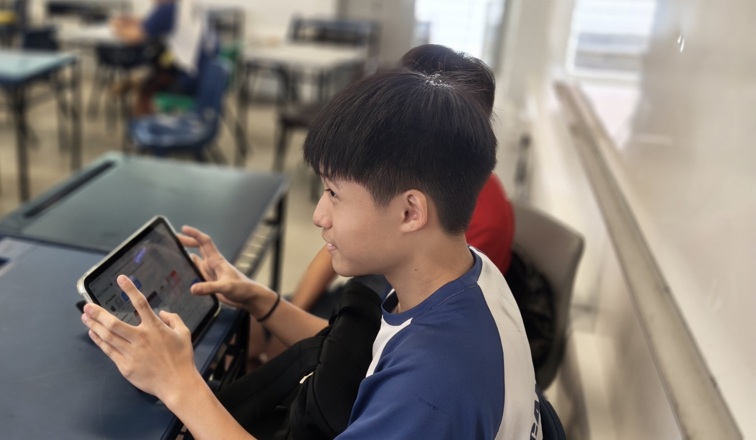 A secondary school boy in a blue and white polo shirt sits at his desk, viewed from the side, holding up a tablet with both hands and looking at its screen with a slight smile. Other students working at their desks are visible in the blurred background.